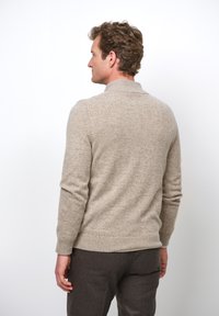 Beige Strickpullover mit hohem Kragen und langen Ärmeln, ausgestattet mit einer strukturierten Oberfläche. Über braunen Hosen getragen, von hinten betrachtet.