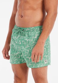 Gröna badshorts med ett upprepande mönster av palmblad, elastisk midja och justerbar snörning. Har en liten logotyp.