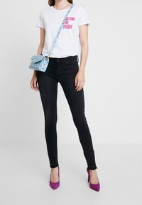 Valkoinen t-paita, jossa on vaaleanpunaista tekstiä, mustat kapeat farkut, sininen käsilaukku, jossa on punaisia sydämiä, ja violetit korkokengät. Yksinkertainen muotoilu ja selkeät linjat.