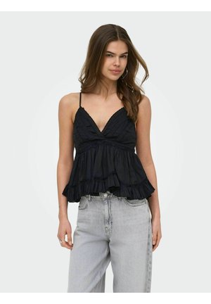 Jeune femme portant un haut noir sans manches à col en V avec des volants, et un jean gris clair taille haute, sur un fond uni.