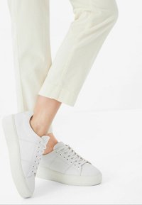Weiße Leder-Sneaker mit strukturiertem Finish, runder Zehenpartie, flachen Schnürsenkeln und einer dicken weißen Sohle, kombiniert mit hellen, gerippten cremefarbenen Hosen.