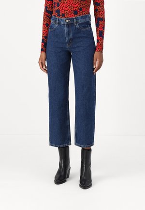Personne vêtue d’un jean bleu taille haute à jambes droites, de bottines noires à talons et d’un haut à manches longues rouge et bleu marine à motif floral.