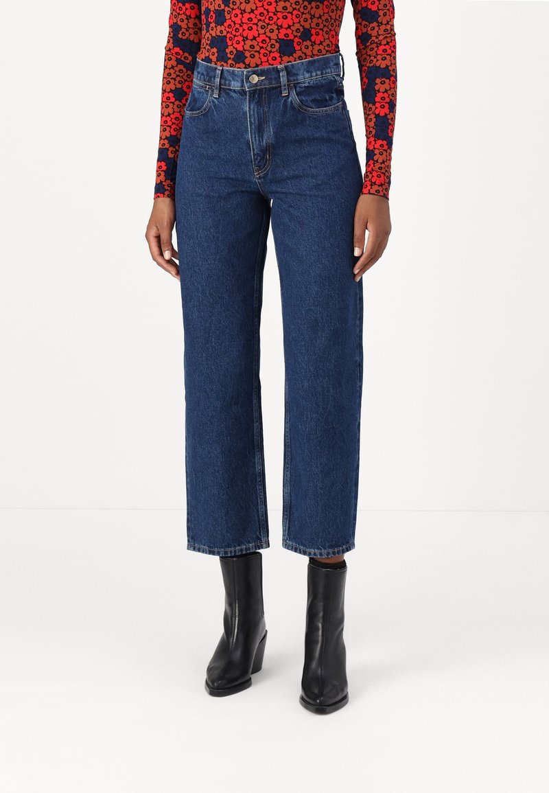Personne vêtue d’un jean bleu taille haute à jambes droites, de bottines noires à talons et d’un haut à manches longues rouge et bleu marine à motif floral.