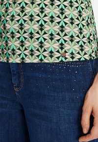 Grün gemusterte Bluse mit floralen Motiven und einem silbernen Logo, kombiniert mit dunkelblauen Jeans, die entlang des Bunds mit silbernen Nieten verziert sind.