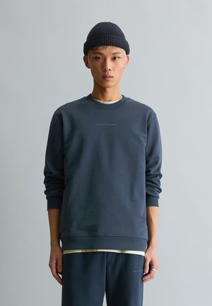 Jeune homme portant un bonnet bleu marine, un sweat-shirt bleu marine et un pantalon assorti, debout devant un fond gris clair uni.