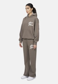 Bruine hoodie en bijpassende joggingbroek gemaakt van een zachte stof, met de tekst "Good for Nothing" in het wit op de voorkant.