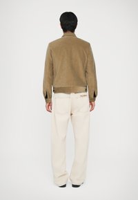 Brūns korduroja žakets ar īsu dizainu, kombinēts ar krēma krāsas platajām biksēm, kurām ir aizmugurējās kabatas un gaiši šuvju punkti. 