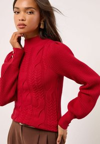 Roter Strickpullover mit gerippter Struktur, der über Zopfmuster, einen hohen Halsausschnitt und puffige Ärmel verfügt, die mit Knopfakzenten gesichert sind.