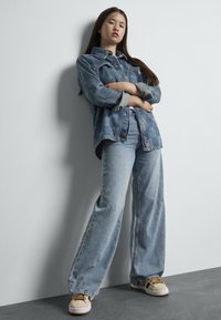 Γυναίκα που στηρίζει την πλάτη της στον τοίχο με τα χέρια σταυρωμένα, φορώντας τζάκετ από καρό τζιν, παντελόνι φαρδιάς γραμμής και μπεζ αθλητικά παπούτσια με κίτρινες λωρίδες.