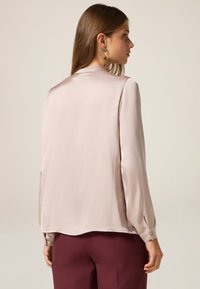 Himmelblaue Satinbluse mit langen Ärmeln, enganliegenden Manschetten mit Knöpfen und einer glatten, glänzenden Textur. Kombiniert mit kastanienbraunen Hosen.