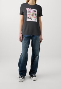 Camiseta de algodón gris con un gráfico de dibujos animados colorido con acentos en rosa y verde. Combinada con unos pantalones vaqueros claros y desgastados y zapatillas negras.