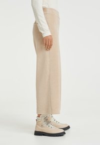 Béžové kordové kalhoty s širokými nohavicemi, které jsou nošeny s světle zbarvenými botami s kovovými prvky a strukturovanou podrážkou. Šňůrky bot jsou uvázané.