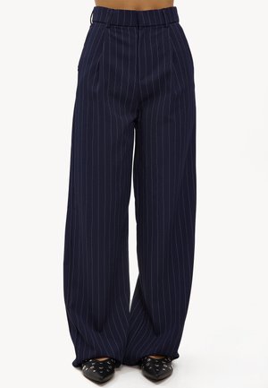Pantalons bleu marine à jambes larges avec des rayures blanches verticales, présentant un devant plissé et une taille haute. Associés à des chaussures noires pointues.