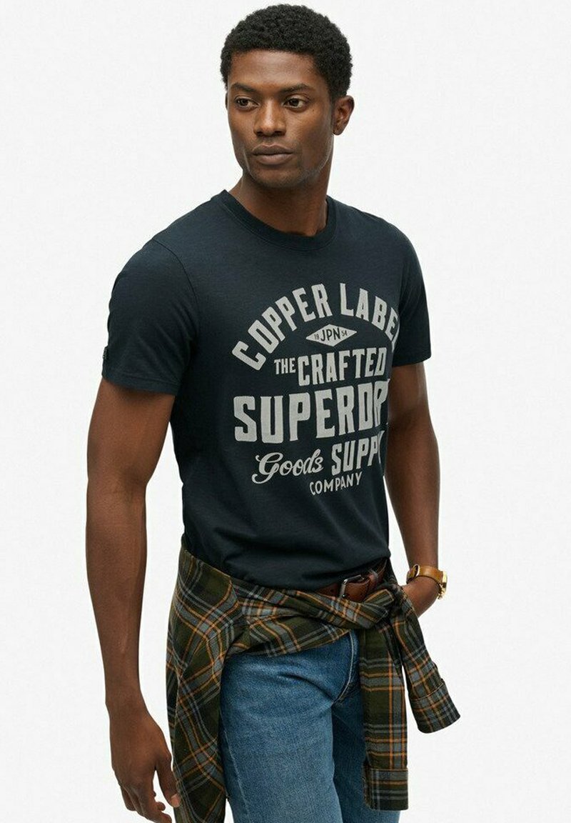 Mand iført sort grafik t-shirt, blå jeans og en ternet skjorte bundet om taljen, ser ud til siden mod hvid baggrund.