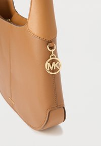 Bolso de piel color canela con colgante de logo MK dorado en el asa, mostrando detalles en piel texturizada y lisa sobre fondo blanco.