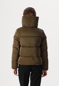 Olivenfarvet dynejakke med ternet mønster, høj krave og quiltet tekstur. Har et figursyet design med markante syninger.