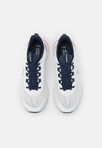 Chaussures de sport blanches avec tige en mesh respirant, lacets bleu marine et accents rouges au talon. Présentent les marques "Ortholite" et "Lacoste Sport".