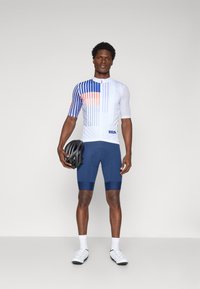 Short-sleeve cycling jersey in white with blue and orange graphic lines, paired with navy shorts, holding a black helmet, model standing.