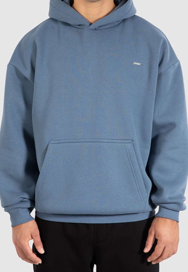 Personne portant un sweat à capuche bleu uni avec une poche avant et un pantalon noir, debout devant un fond clair uni.