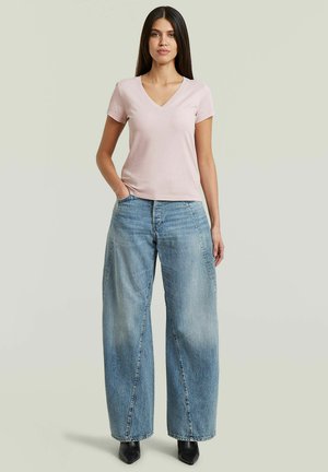 Vrouw met steil haar, staand, gekleed in een lichtroze T-shirt met V-hals, wijde lichtblauwe spijkerbroek en zwarte puntige laarzen.