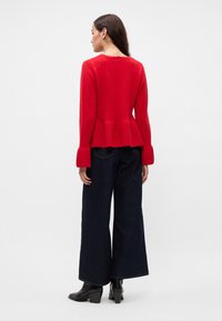 Pulover roșu tricotat cu un design peplum și mâneci largi, purtat cu blugi negri cu croială largă și botine negre.