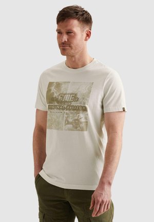 Mann trägt weißes T-Shirt mit Grafik und dem Schriftzug „Original Aviator“ und grüne Cargohosen, steht vor einem einfarbigen hellen Hintergrund.