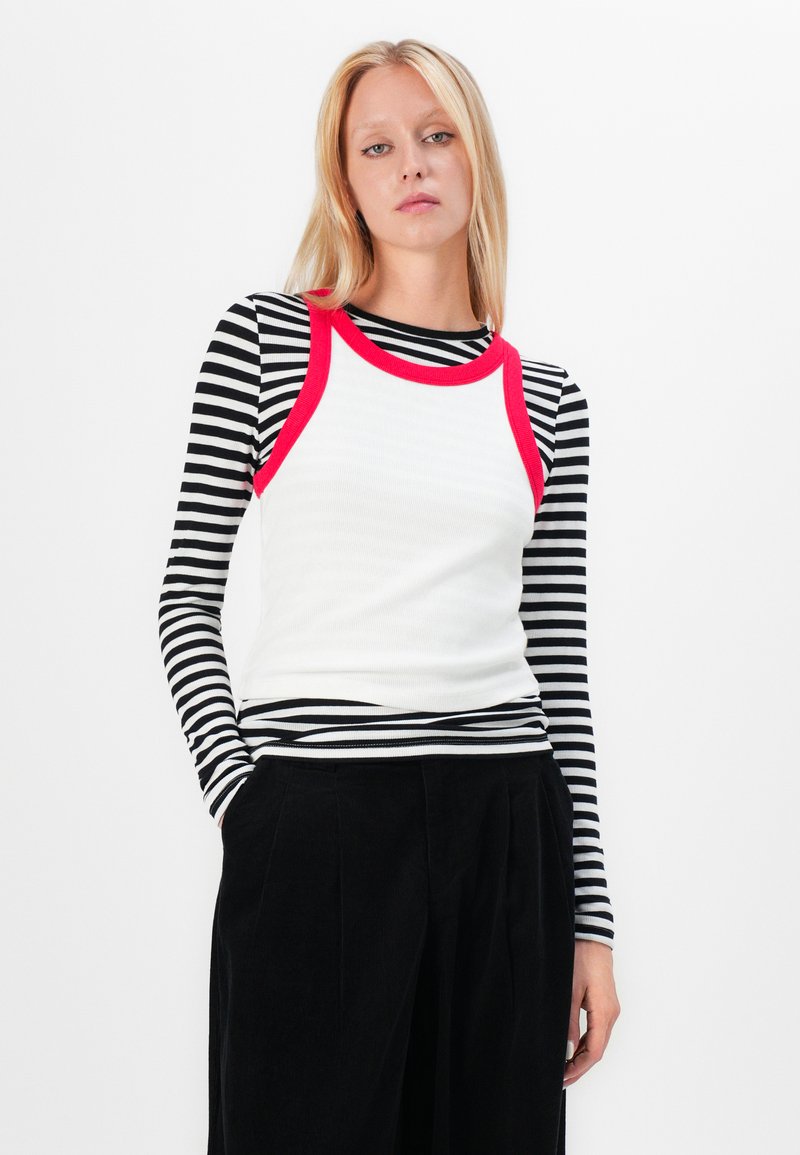 Weißes Tanktop mit rotem Rand über einem schwarz-weiß gestreiften Langarmshirt, kombiniert mit schwarzer, faltiger Hose. Glatte, anliegende Texturen.