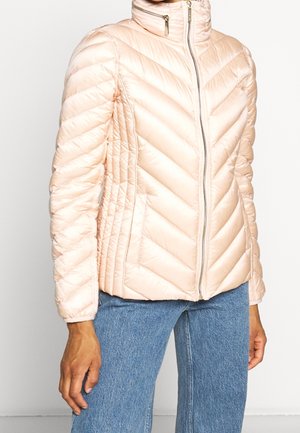Veste matelassée rose clair avec col montant et fermeture éclair à l'avant, portée avec un jean bleu, montrée du cou aux hanches avec les mains détendues.