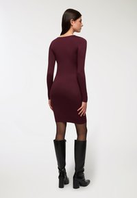 Burgundowa dopasowana sukienka z długimi rękawami, o długości do połowy uda, zestawiona z czarnymi botkami do kolan i przezroczystymi czarnymi rajstopami. Gładka tekstura.