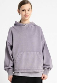 Młoda osoba w jasnofioletowej, luźnej bluzie z kapturem z przednią kieszenią i czarnych spodniach, stojąca na jednolitym białym tle.