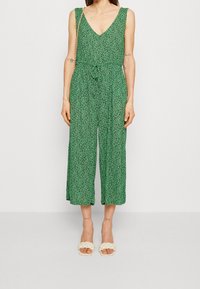 Groene mouwloze jumpsuit met een V-hals, cinched taille en cropped wijde benen met een subtiel patroon. Gecombineerd met crèmekleurige gevlochten sandaaltjes.