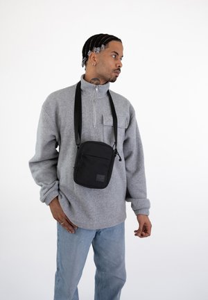 Black crossbody bag made of smooth fabric, featuring a rectangular shape, adjustable strap, and a small logo on the front. Worn over a gray fleece pullover.