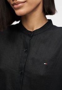 Γυναίκα που φοράει μαύρο πουκάμισο με μικρό ορθογώνιο λογότυπο σε κόκκινο, άσπρο και μπλε στην αριστερή πλευρά του στήθους.