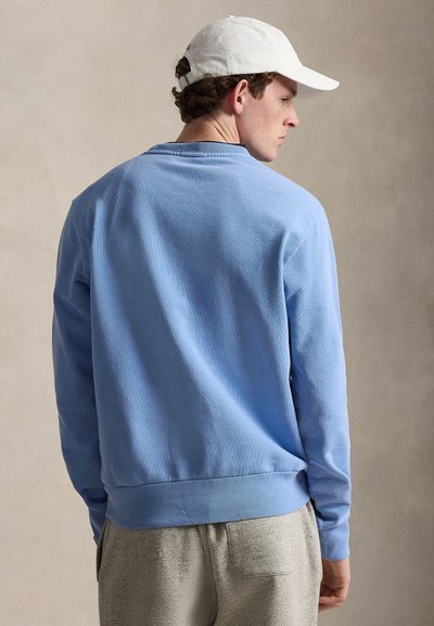 Άνδρας που φοράει λευκό καπέλο, ανοιχτό μπλε φούτερ και γκρι φόρμα, στραμμένος πλάγια σε απλό μπεζ φόντο.