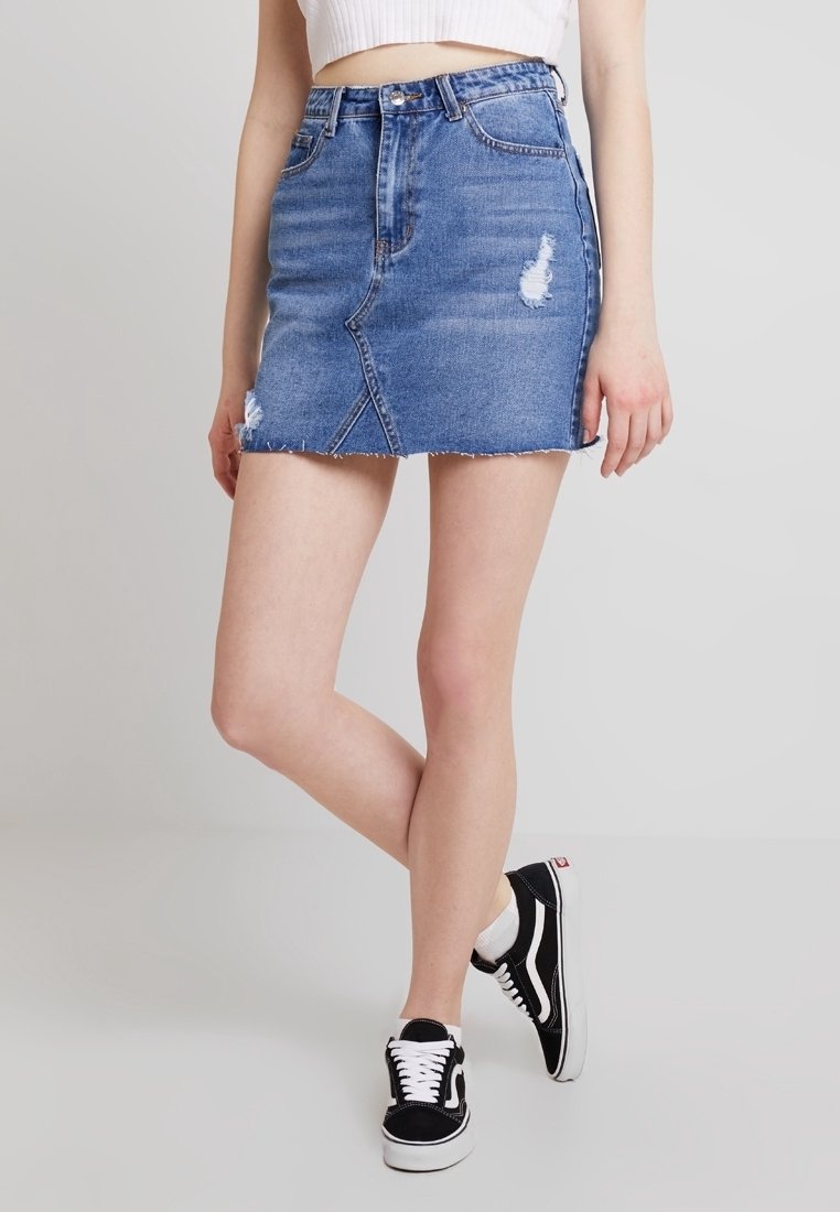 Jupe en jean mini avec ourlet effiloché, couleur bleu moyen, poches avant et détails abîmés. Associée à des baskets noires et blanches.