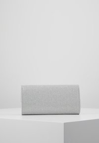 Petit sac pochette rectangulaire argenté pailleté reposant sur un socle blanc, sur un fond gris clair uni.