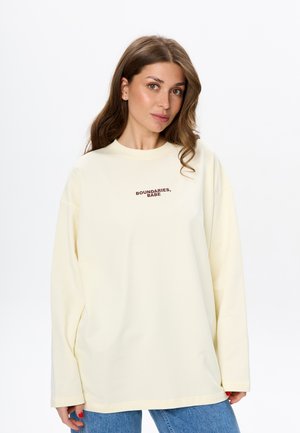 Crème oversized sweatshirt met lange mouwen, ronde halslijn en donkerbruine tekst "BOUNDARIES, BABE" op de borst. Draagt met blauwe jeans.
