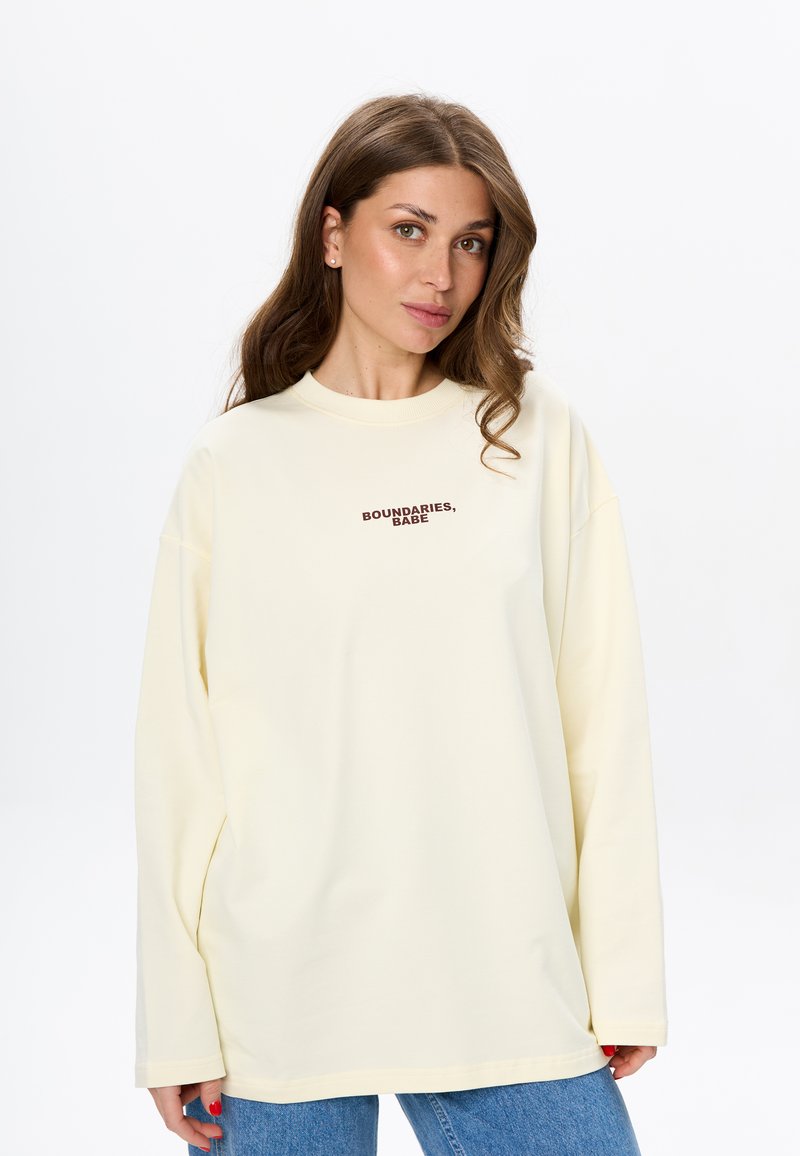 Cream oversized sweatshirt with long sleeves, round neckline, and dark brown text "BOUNDARIES, BABE" on the chest. Worn with blue jeans.