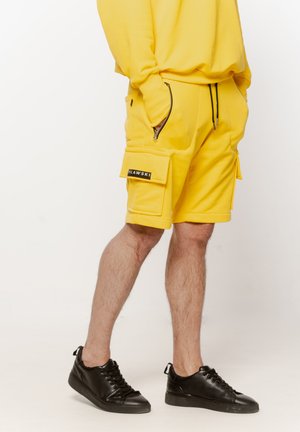 Mężczyzna w żółtych szortach cargo z kieszeniami na zamek i klapą, czarnych trampkach oraz pasującej żółtej bluzie, stojący na białym tle.