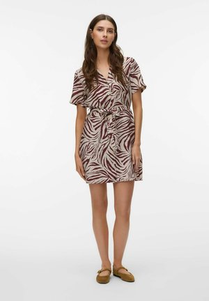 Vrouw draagt een kortärmelig, ceintuurlinnen jurk met kastanjebruine en beige zebraprinten, staand met ontspannen handen, draagt bruine platte schoenen.