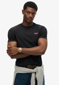 Zwarte korte mouw t-shirt met roze logo, gecombineerd met een grijze trui om de taille geknoopt. Verfraaid met een gouden horloge.