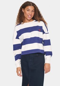 Gekürzter Sweatshirt in Creme mit marineblauen Streifen. Verfügt �über ein rotes Logodetail, lange Ärmel und eine entspannte Passform. Kombiniert mit einem dunklen Rock.