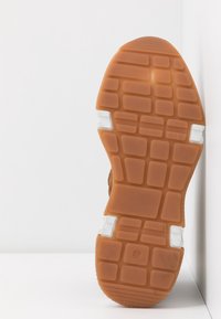 Semelle de chaussure avec un motif de bande de roulement en caoutchouc brun et des accents blancs, appuyée contre un angle blanc entre mur et sol.