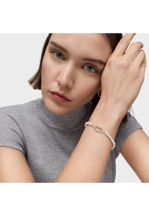 Kobieta o krótkich brązowych włosach, ubrana w szary golf z krótkim rękawem, pokazuje na uniesionym nadgarstku perłową bransoletkę z srebrną zapinką.