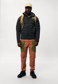 Giacca a vento nera, pantaloni arancioni e verdi con tasche, scarpe nere, cappello giallo e uno zaino giallo con un moschettone attaccato.