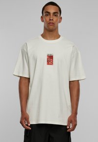 Kremfarget oversized bomull t-skjorte med et kvadratisk grafisk lapp som har røde blomster og teksten "PEACE NO WAR." Korte ermer, rund hals.