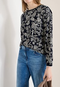 Femme portant un haut noir à manches longues avec un motif floral blanc et un jean bleu, tenant un vêtement marron dans sa main droite.