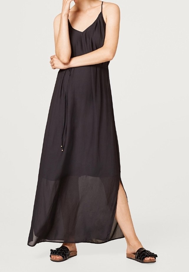 Femme portant une robe maxi noire sans manches avec des bretelles spaghetti et des sandales noires à volants, debout avec une jambe exposée.