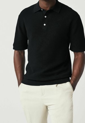 Czarna, teksturowana koszulka polo z krótkim rękawem, z trzema guzikami z przodu i prążkowanym kołnierzykiem, zestawiona z jasnobeżowymi spodniami.