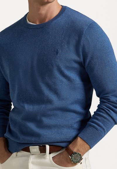 Blauwe trui met een gebreide textuur, ronde halslijn en geborduurd logo op de borst. Gepaard met een bruine riem en een horloge om de pols.